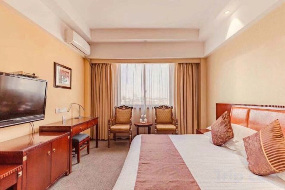 A hotel room with a bed, a desk with a television, two chairs, and a window with curtains.