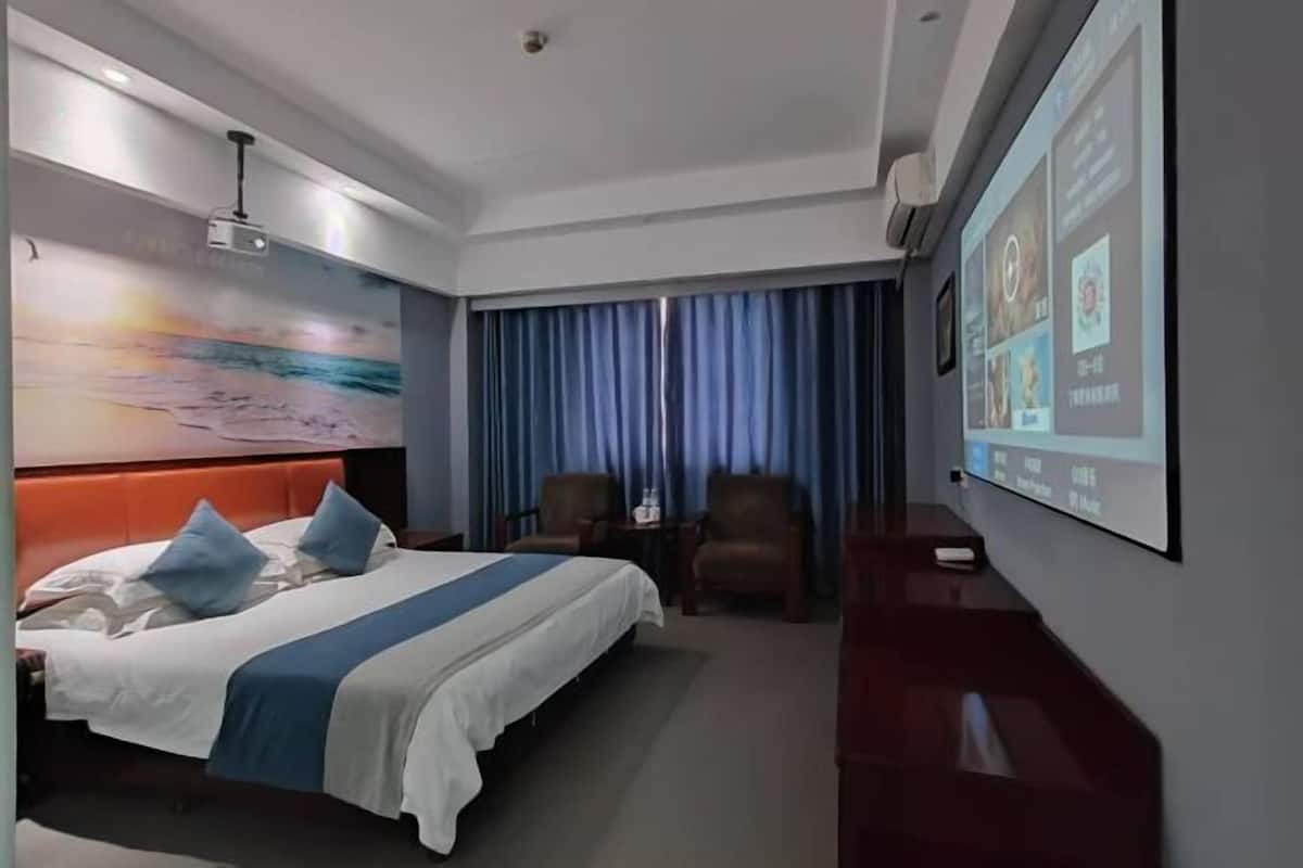 A hotel room with a bed, a desk, a chair, a TV screen, and a painting on the wall.