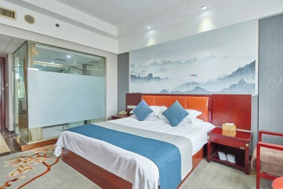 A hotel room with a large bed, a nightstand, a red headboard, a wall painting, and a glass partition.