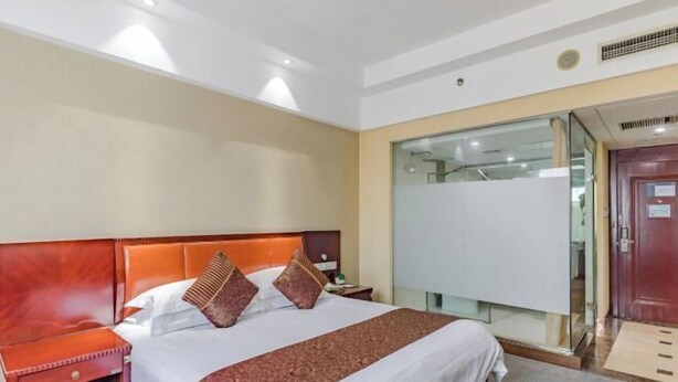 Una habitación de hotel con una cama grande, una mesita de noche de madera, una partición de vidrio y una puerta.