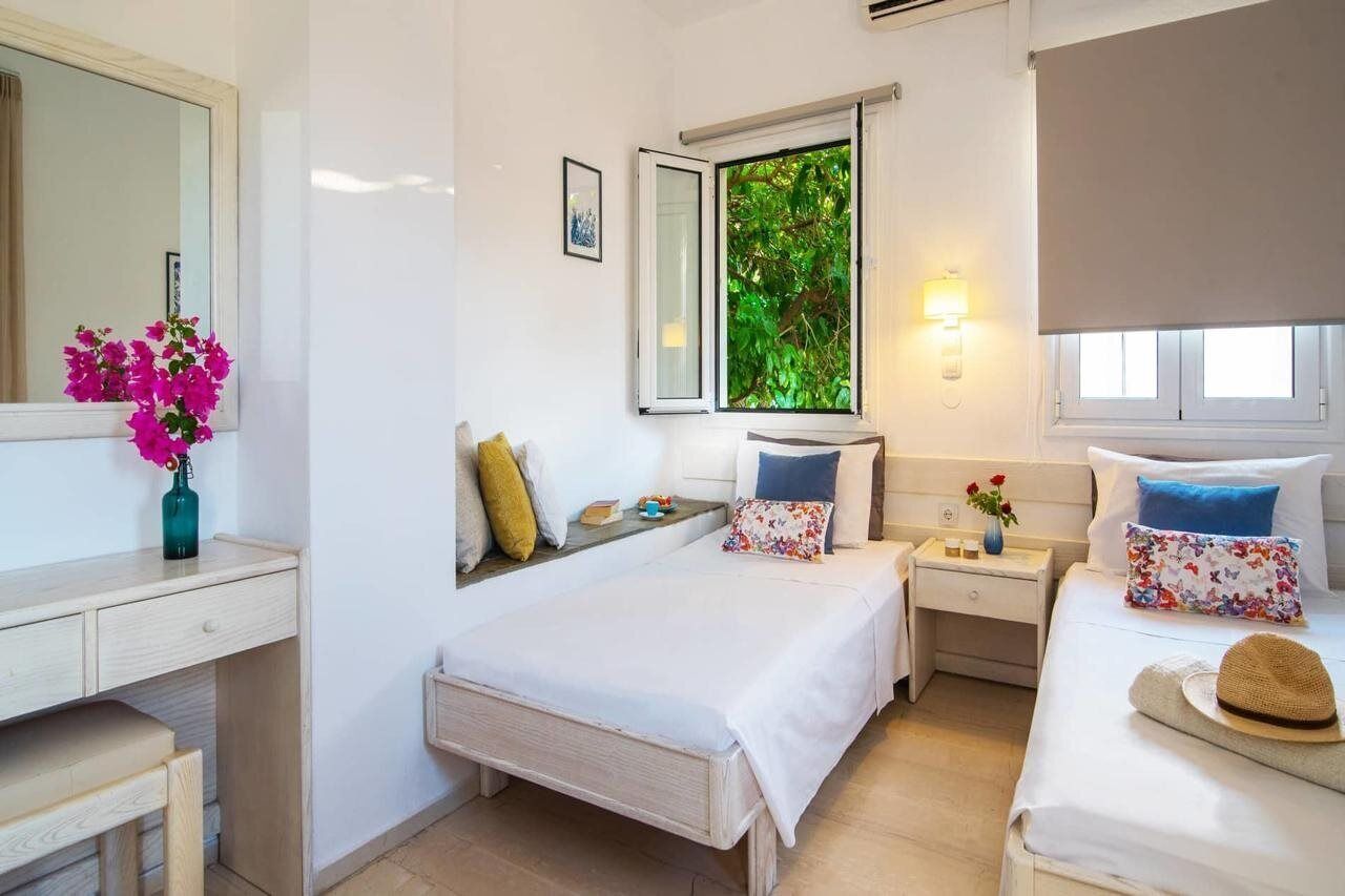 A bedroom with two beds, a desk, and a window with a view of greenery.