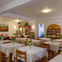 Un restaurante con varias mesas, cada una con mantel blanco y un jarrón de flores. Hay un refrigerador de Coca-Cola y una exhibición de tarros de miel al fondo.