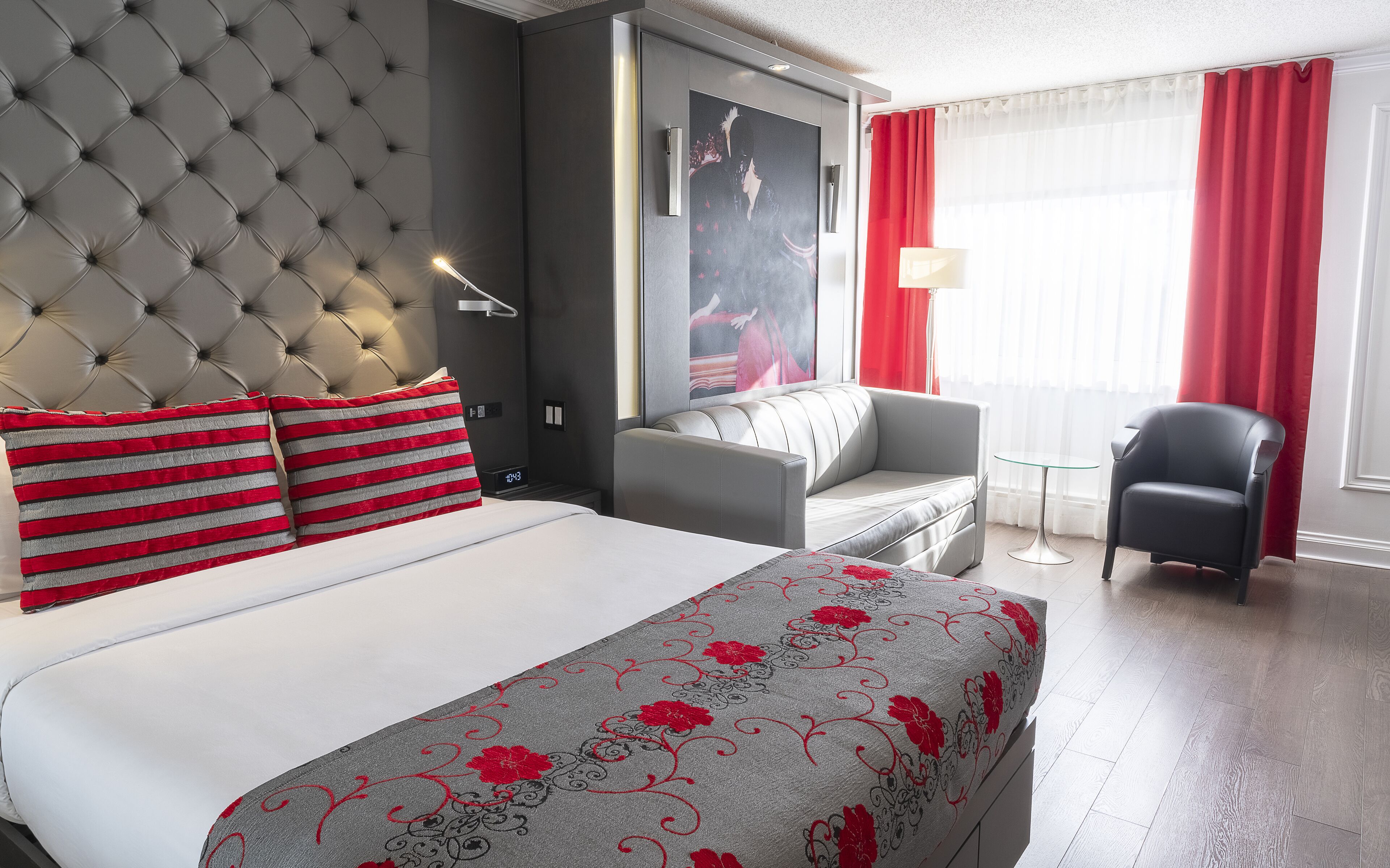 Quarto de hotel com cabeceira estofada, cama com almofadas listradas, sofá cinza, mesa redonda pequena e poltrona preta.