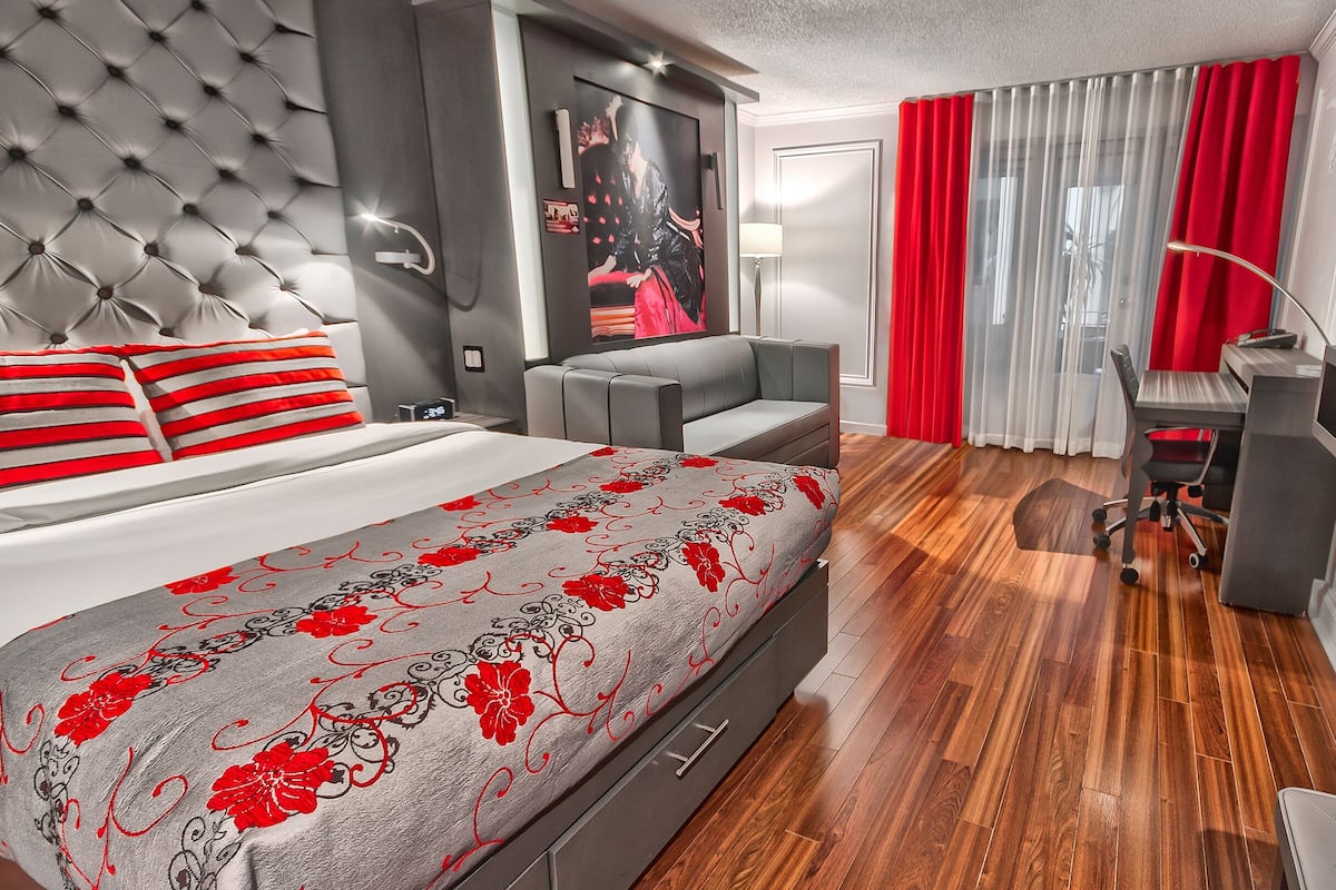 Habitación de hotel moderna con cabecera capitonada, cama con almohadas a rayas rojas y blancas, sofá gris, escritorio con silla y televisor.
