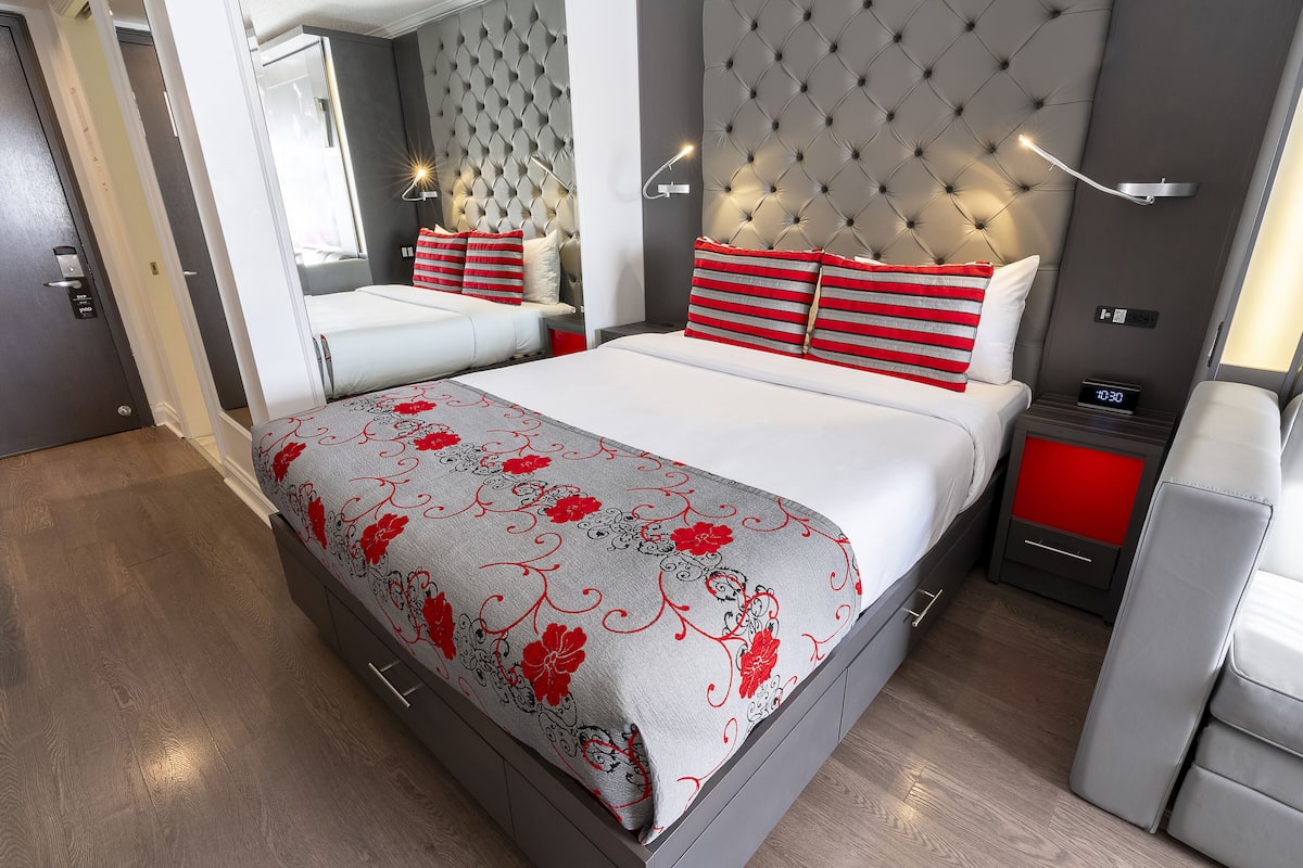 Un dormitorio moderno con cabecera capitonada, una cama con almohadas a rayas rojas y blancas, una mesita de noche con un reloj y un espejo sobre la cama.