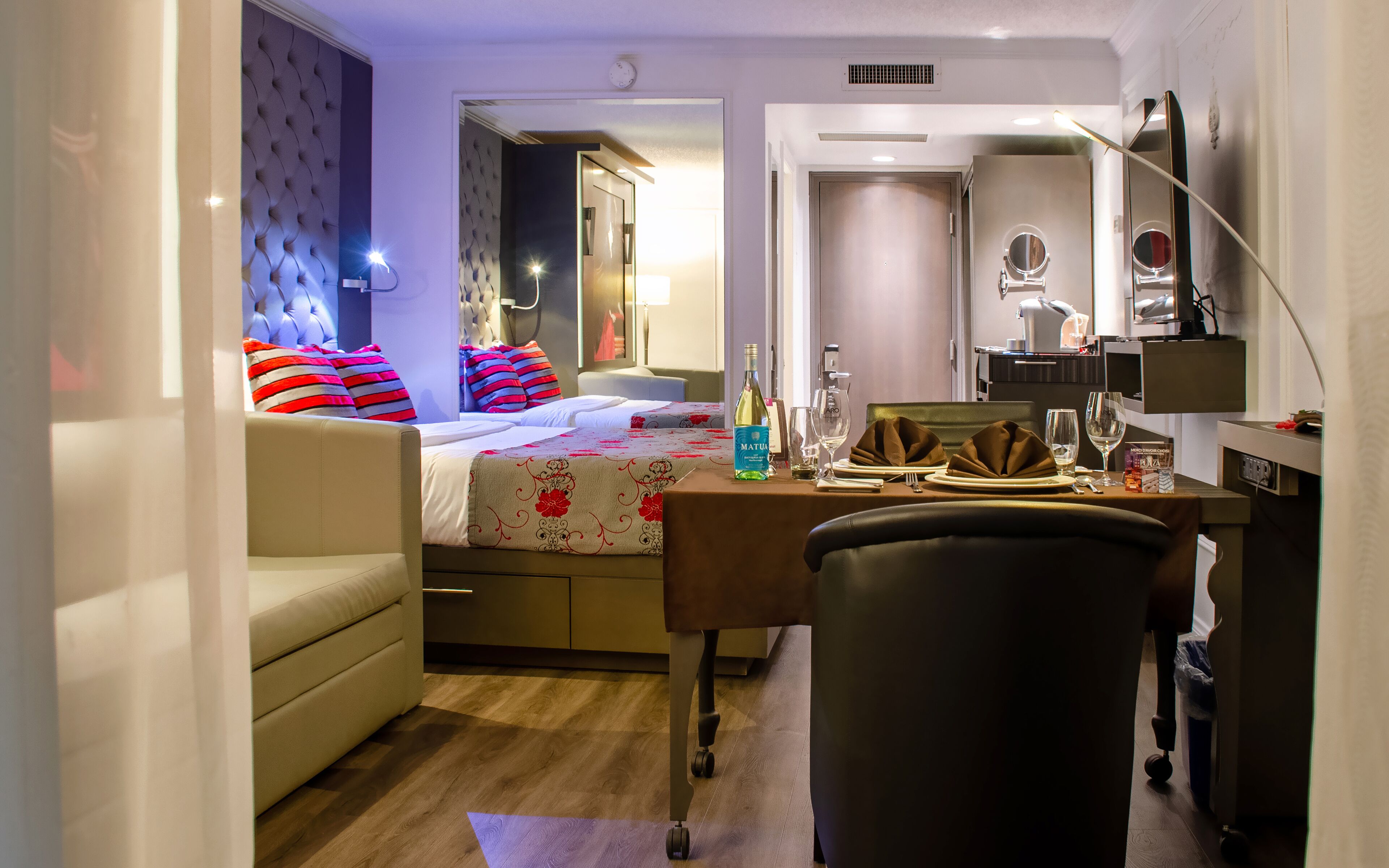 A hotel room with a bed, a desk, a chair, a minibar, and a bathroom visible through an open door.