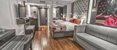 A modern hotel room with a wooden floor, a grey sofa, a bed with striped pillows, and a large mirror.