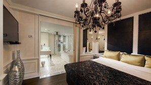 A bedroom with a large bed, a chandelier, a mirror, and a view into the bathroom.