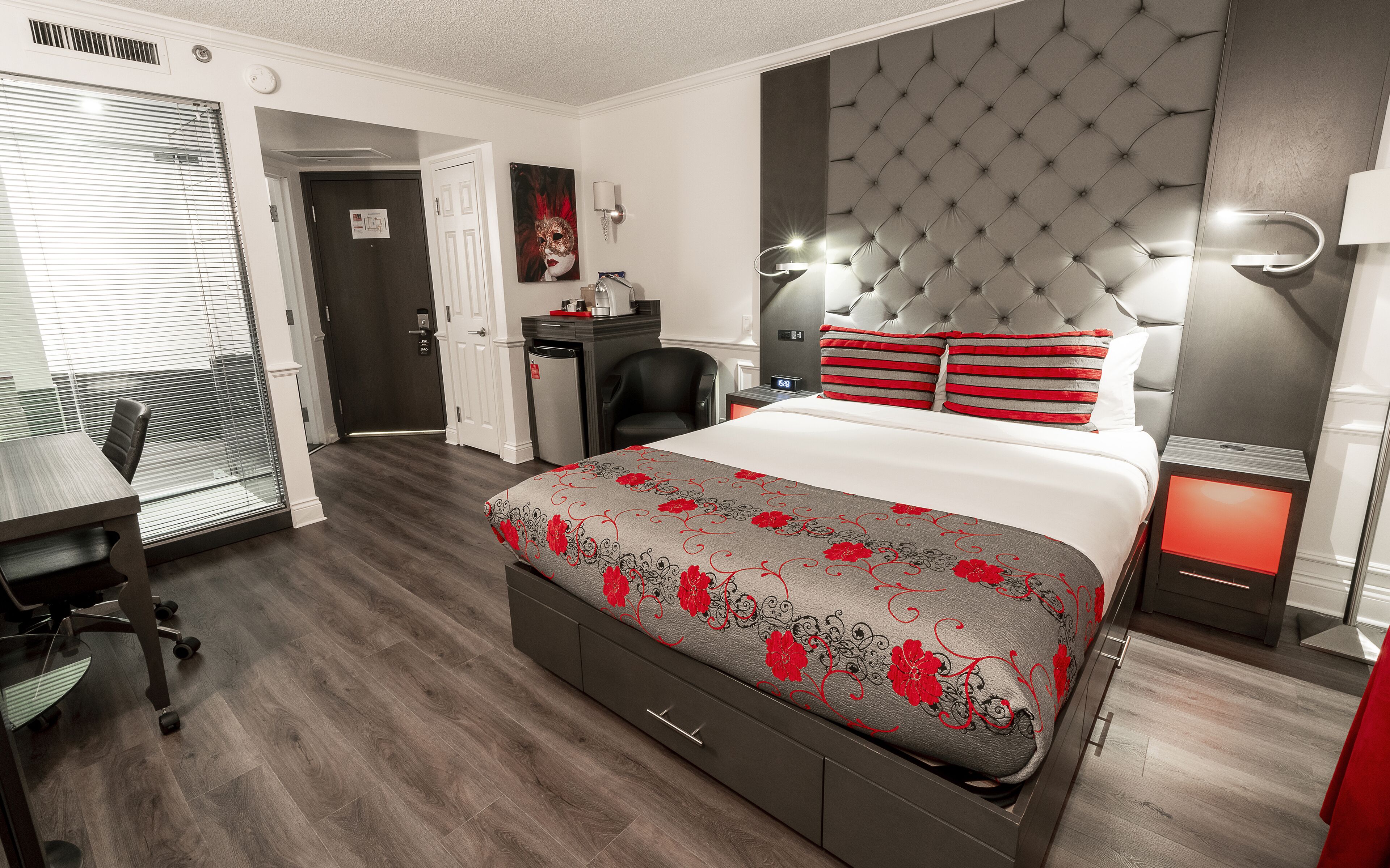 A hotel room with a bed, a desk, a chair, a wardrobe, and a nightstand with a lamp.