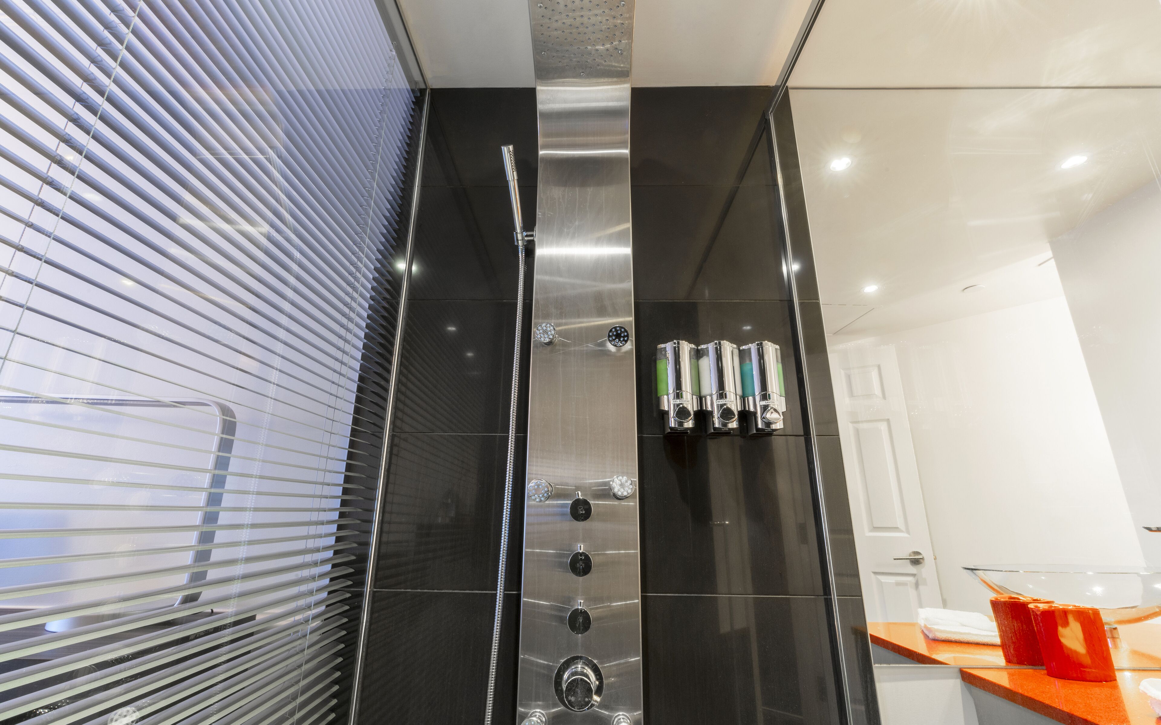 A modern bathroom with a black tiled wall, a shower with multiple controls, and a window with blinds.