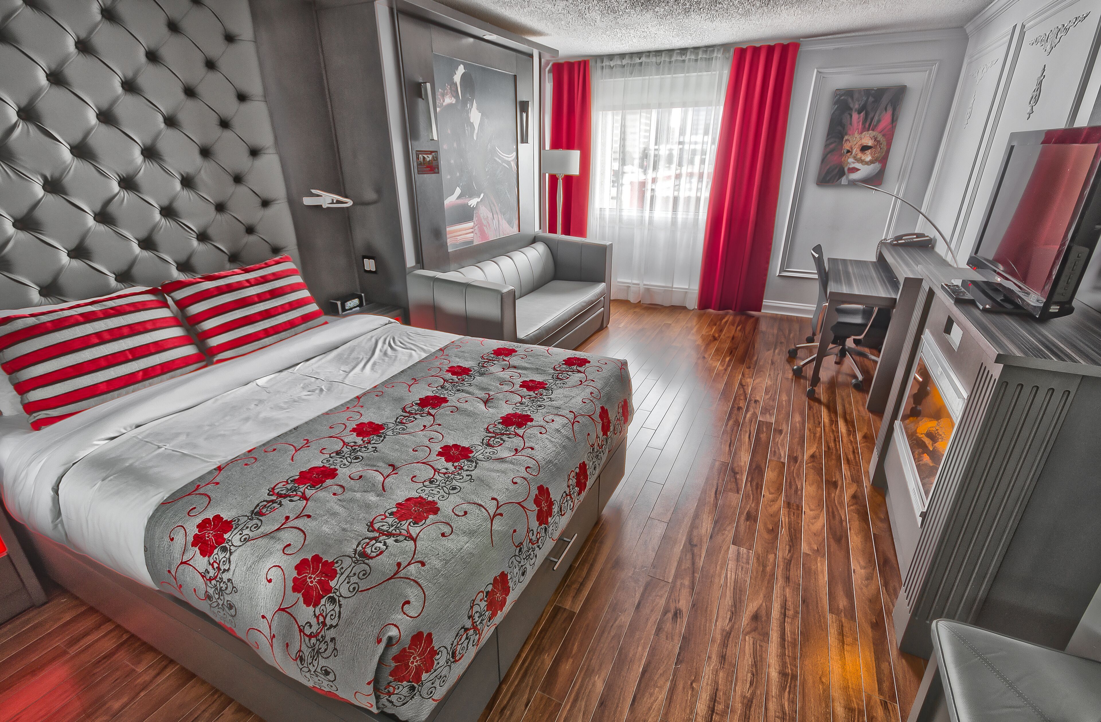Una habitación de hotel moderna con una tapicería capitonada en la cabecera, un sofá gris, un escritorio con una computadora y una cama con ropa de cama floral en tonos rojos y grises.