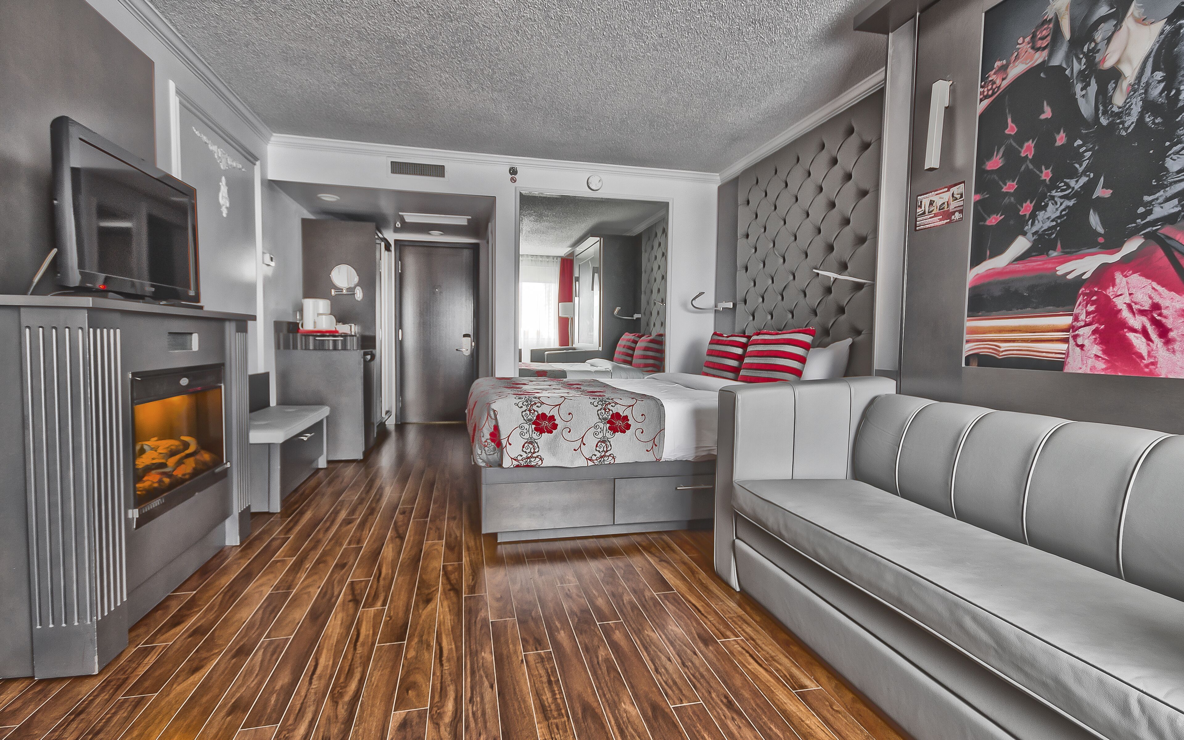 A modern hotel room with a sofa, a bed with red and white striped pillows, a television, and a fireplace.