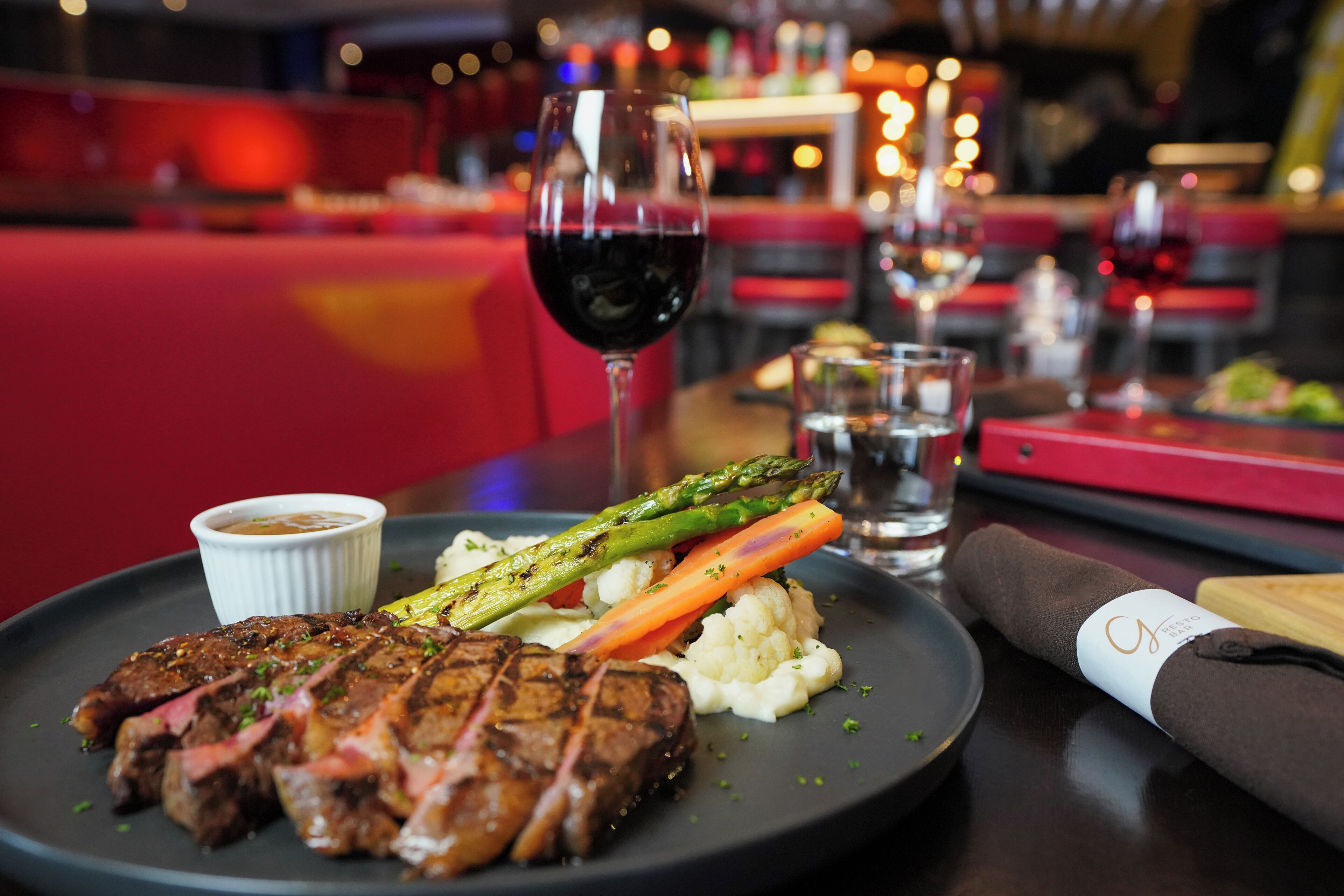 A plate with steak, asparagus, and carrots, a glass of red wine, and a small bowl of sauce.