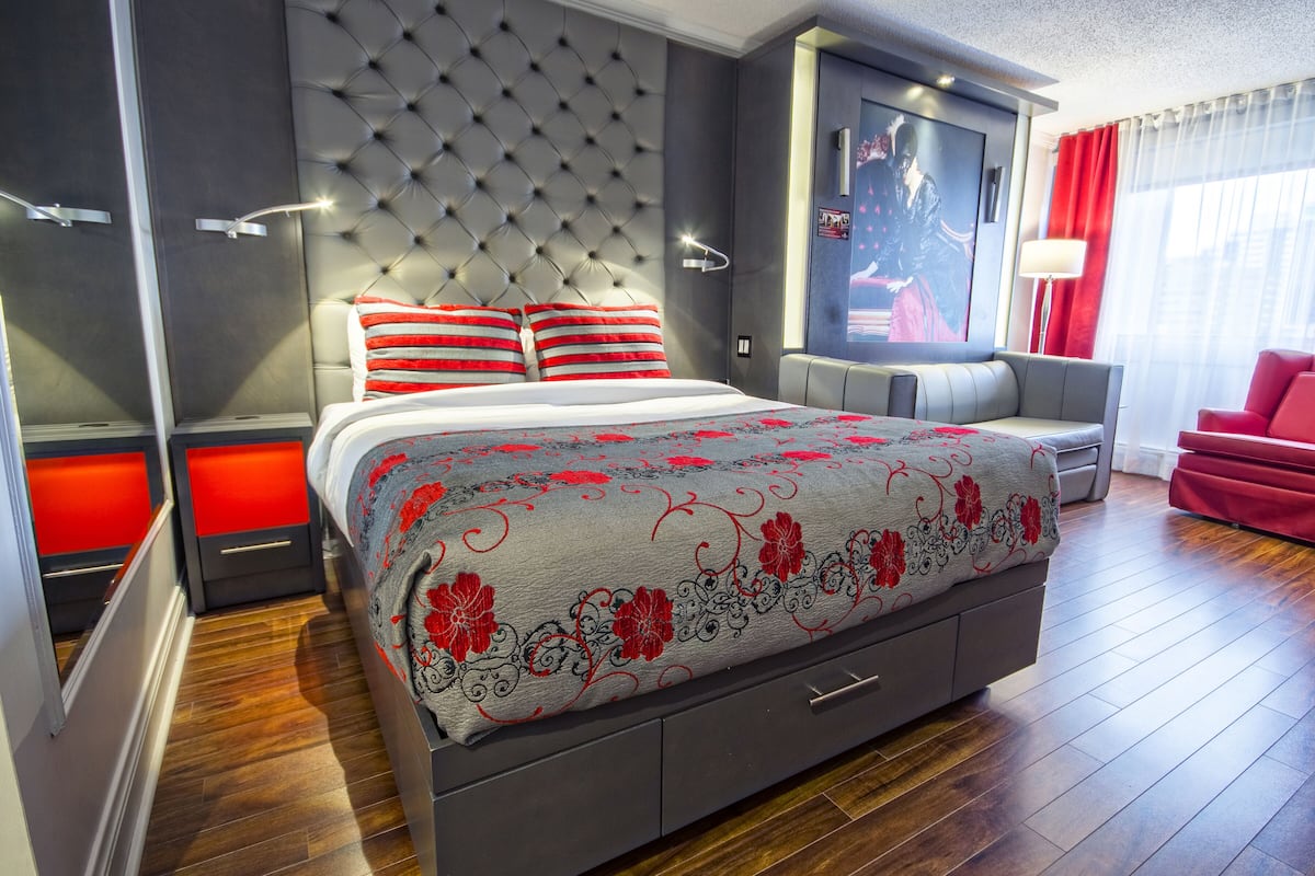 Una habitación de hotel moderna con una cama grande, cabecera tapizada en gris, almohadas a rayas rojas y blancas, un sofá gris y un sillón rojo.