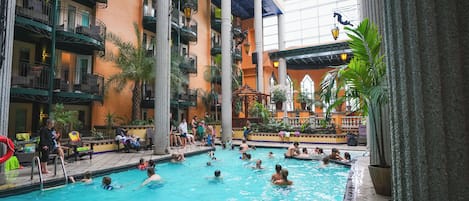 An indoor swimming pool with multiple levels of balconies, surrounded by tropical plants and a covered upper area with large windows.
