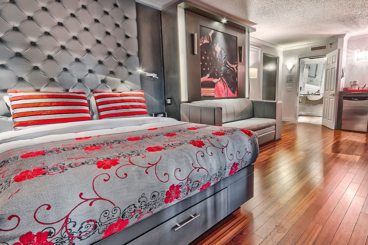 Un dormitorio moderno con cabecera capitonada, una cama grande con ropa de cama roja y gris, un sofá gris y suelo de madera.