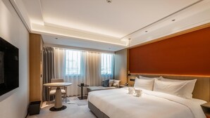Une chambre d’hôtel comprenant un lit, une table de chevet, une petite table avec des chaises, un téléviseur fixé au mur et une fenêtre avec des rideaux.