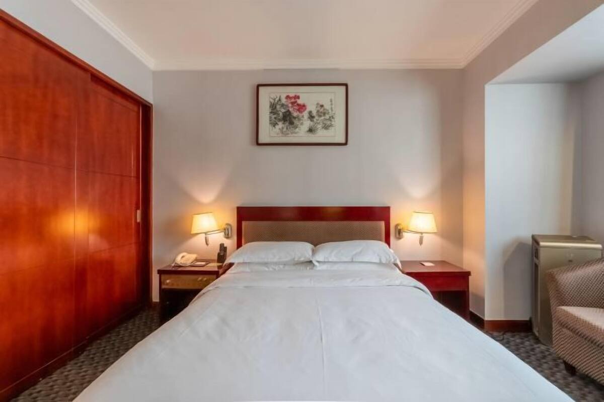 A hotel room with a large bed, two bedside lamps, a wooden wardrobe, a framed artwork on the wall, and a chair.