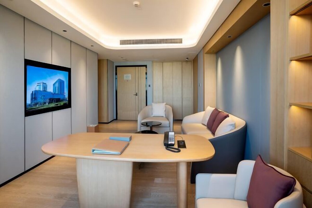 Une chambre d’hôtel moderne avec un bureau en bois aux lignes courbes, deux fauteuils blancs, un téléviseur à écran plat fixé au mur et une porte donnant sur une autre pièce.