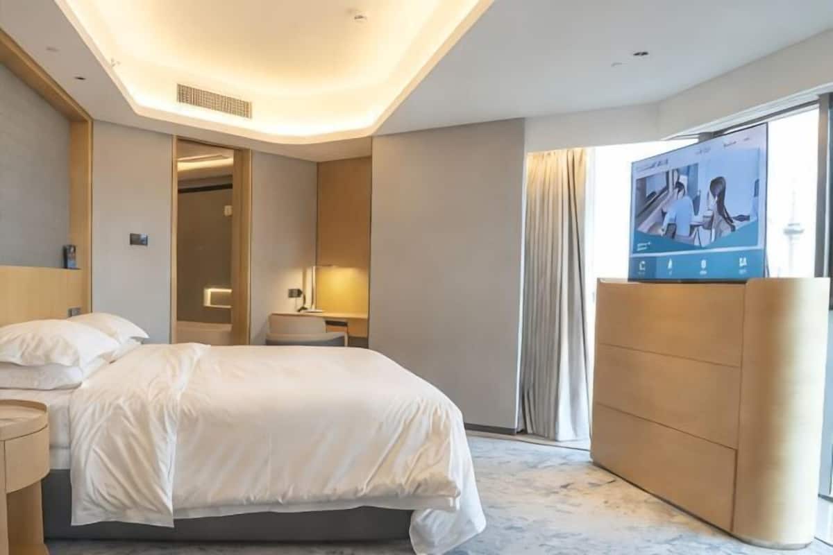 A modern hotel room with a large bed, a TV on a stand, a bedside table, and a view of the outside.