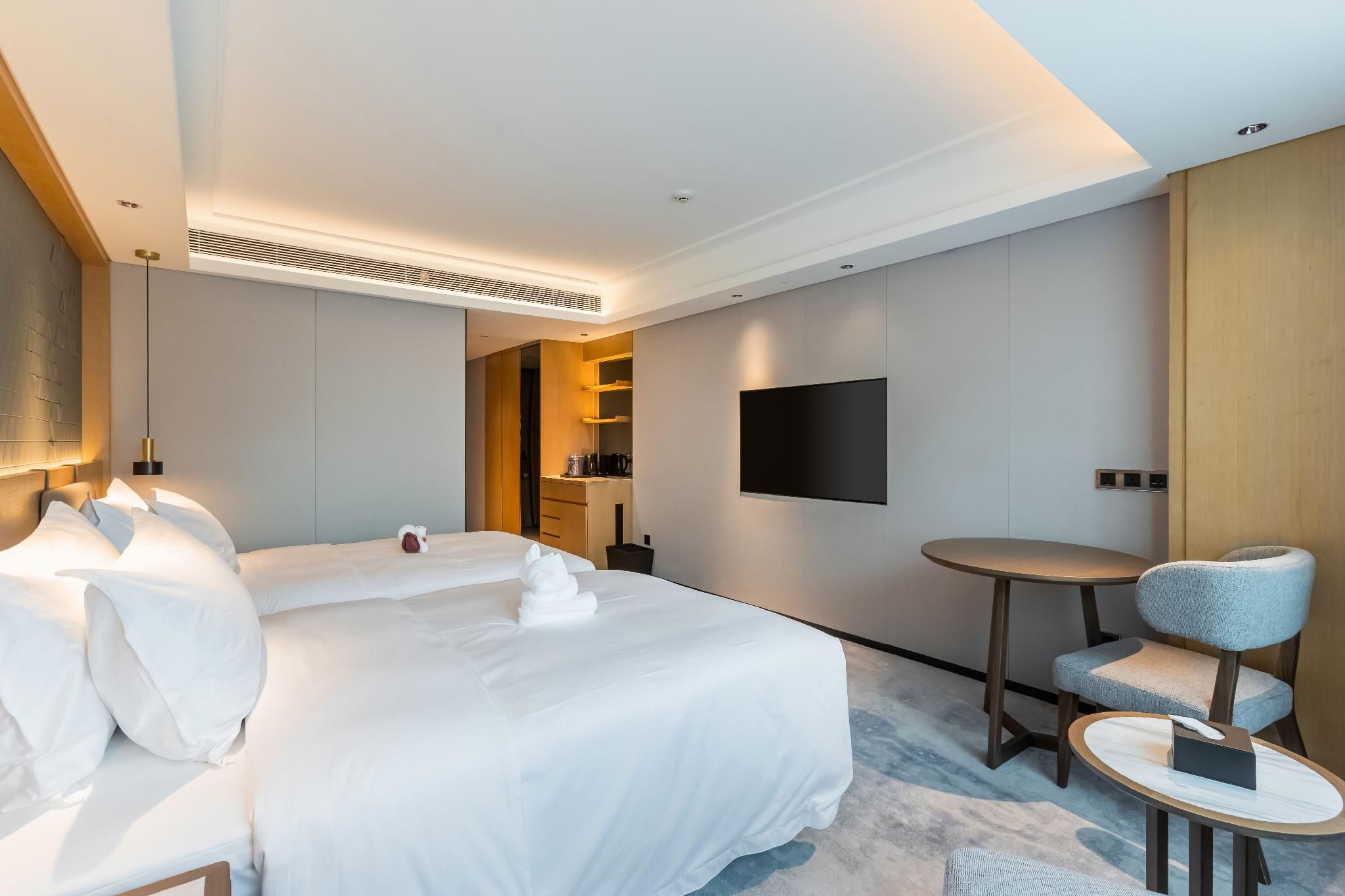 A modern hotel room with a large bed, bedside tables, a flat-screen TV mounted on the wall, a small round table with a chair, and a built-in kitchen area.