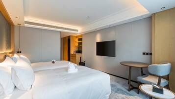 A modern hotel room with a large bed, bedside tables, a flat-screen TV mounted on the wall, a small round table with a chair, and a built-in kitchen area.