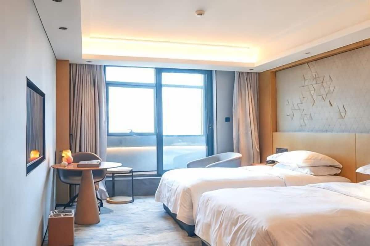 A hotel room with two beds, a desk, a chair, and a large window.