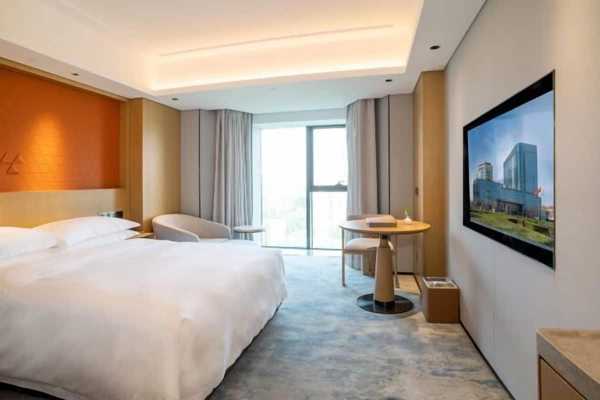 A hotel room with a large bed, a sitting area with a table and chairs, a large window with curtains, and a painting on the wall.