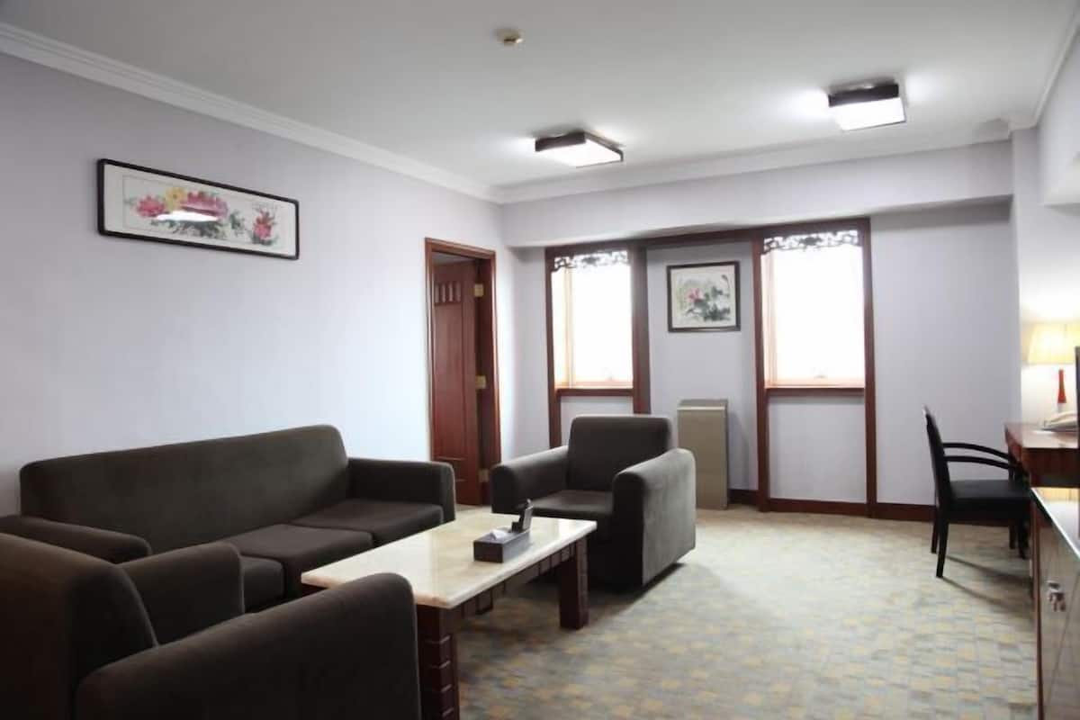 Ein Zimmer mit einer Couch, Sesseln, einem kleinen Tisch und einem Schreibtisch mit Stuhl. Zwei Fenster mit Vorhängen, eine Tür und ein gerahmtes Bild sind an den Wänden zu sehen.