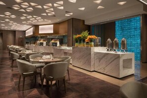A modern hotel lobby with a marble reception desk, dining area with round tables and chairs, and a decorative wall with a blue mosaic.