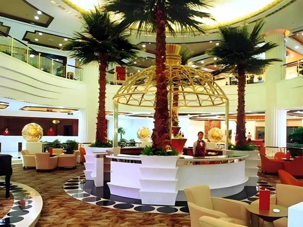 A lobby with a central reception desk, palm trees, and a modern design.
