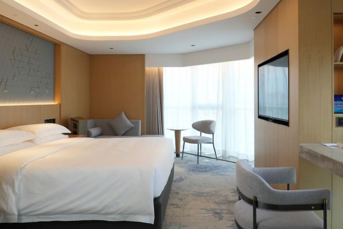 Une chambre d’hôtel avec un grand lit, un bureau, une chaise, une télévision et une fenêtre avec des rideaux.