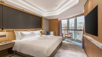 A modern hotel room with a large bed, a flat-screen TV, a city view, and a balcony.