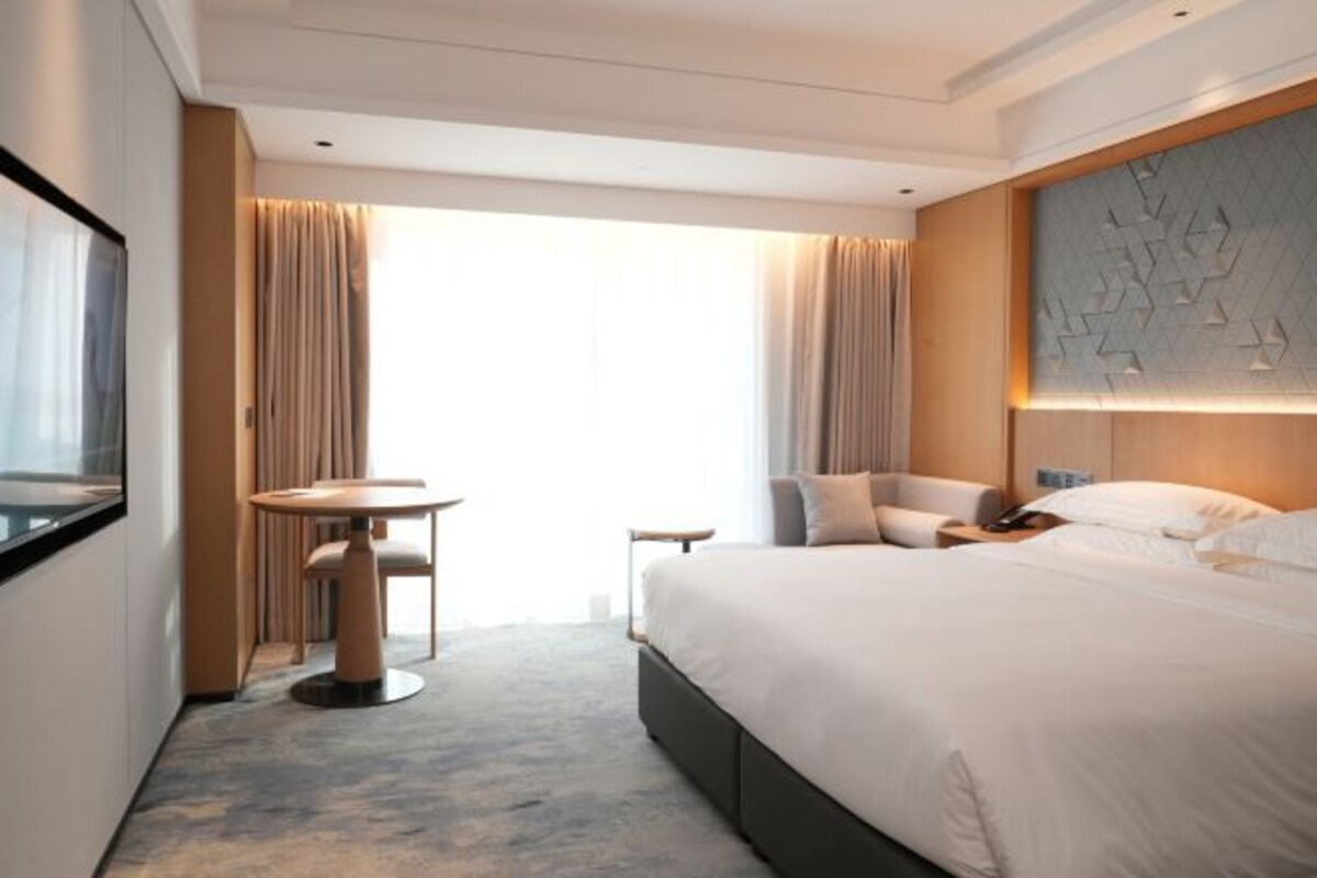 Une chambre d’hôtel avec un lit, une table ronde, une chaise et une grande fenêtre avec des rideaux.