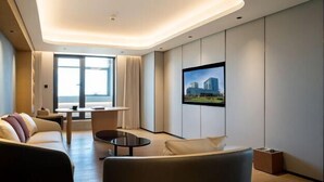 Ein modernes Hotelzimmer mit einer Couch, einem kleinen Tisch, einem an der Wand montierten Fernseher und einem großen Fenster mit Blick auf die Stadt.