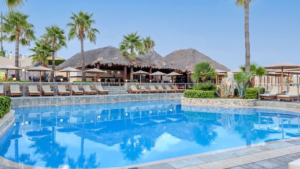 A swimming pool with lounge chairs and umbrellas, surrounded by palm trees and thatched-roof structures. - Eurohotel Theo Hotel (Chania)