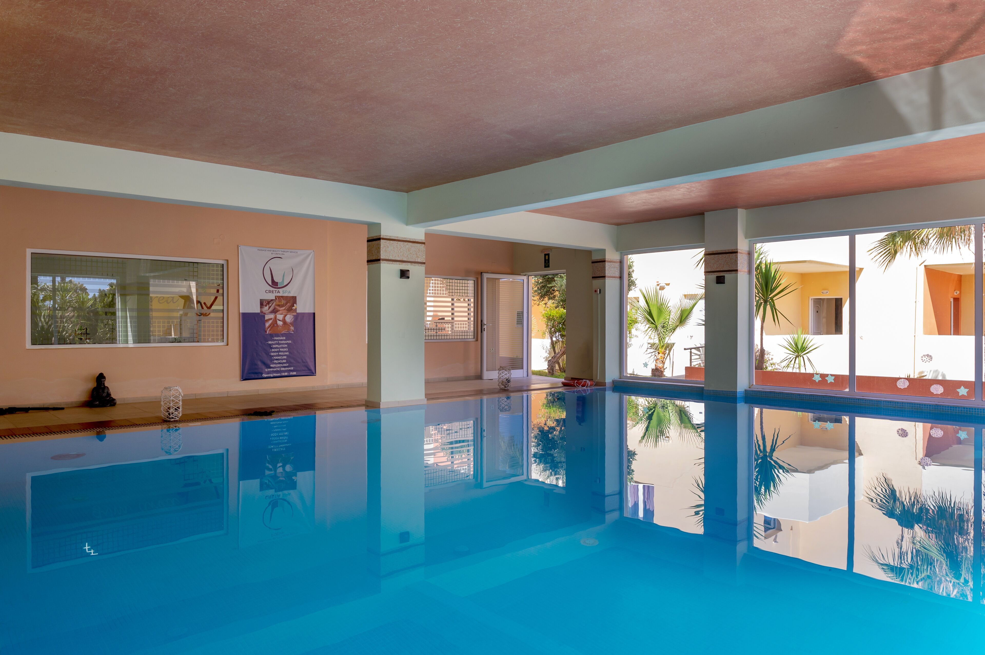 A swimming pool with clear water, a ceiling with a textured finish, and a view of palm trees outside.
