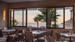 A restaurant with tables and chairs arranged for dining, large windows offering a view of the sea and palm trees. - Eurohotel Theo Hotel (Chania)