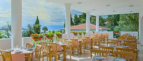 A restaurant with tables set for dining, white columns, and a view of the sea.
