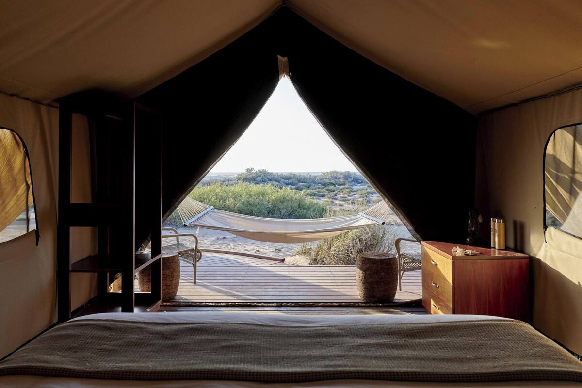 Een tent met een bed, een houten bureau en uitzicht op het strand en de duinen.