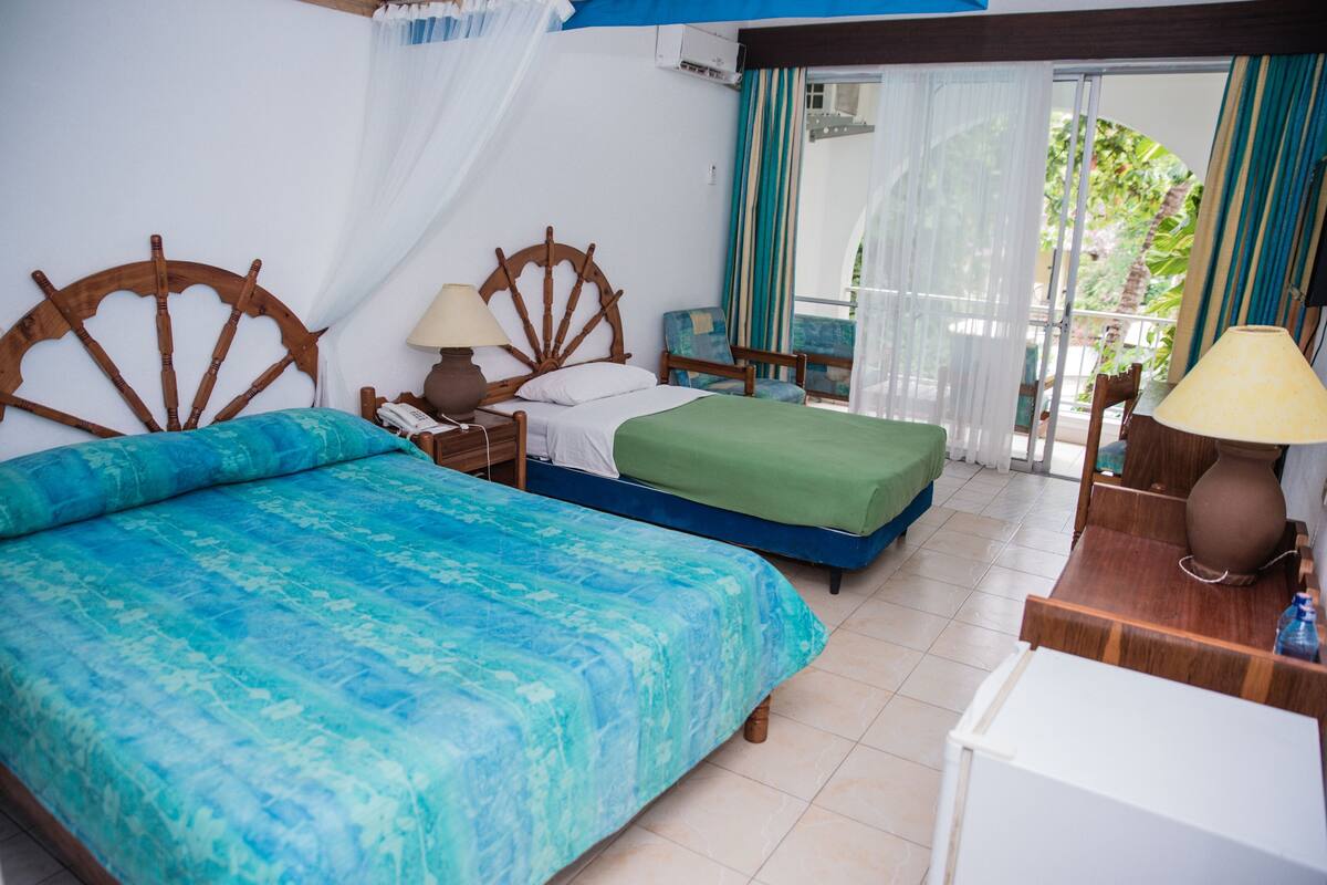 A hotel room with two beds, a small table, a nightstand, and a view of the outdoors.