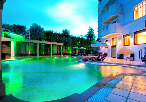 A swimming pool with lounge chairs and umbrellas, surrounded by a building with balconies and a covered area with a sign. - Howard Plaza The Fern, Agra (Agra)