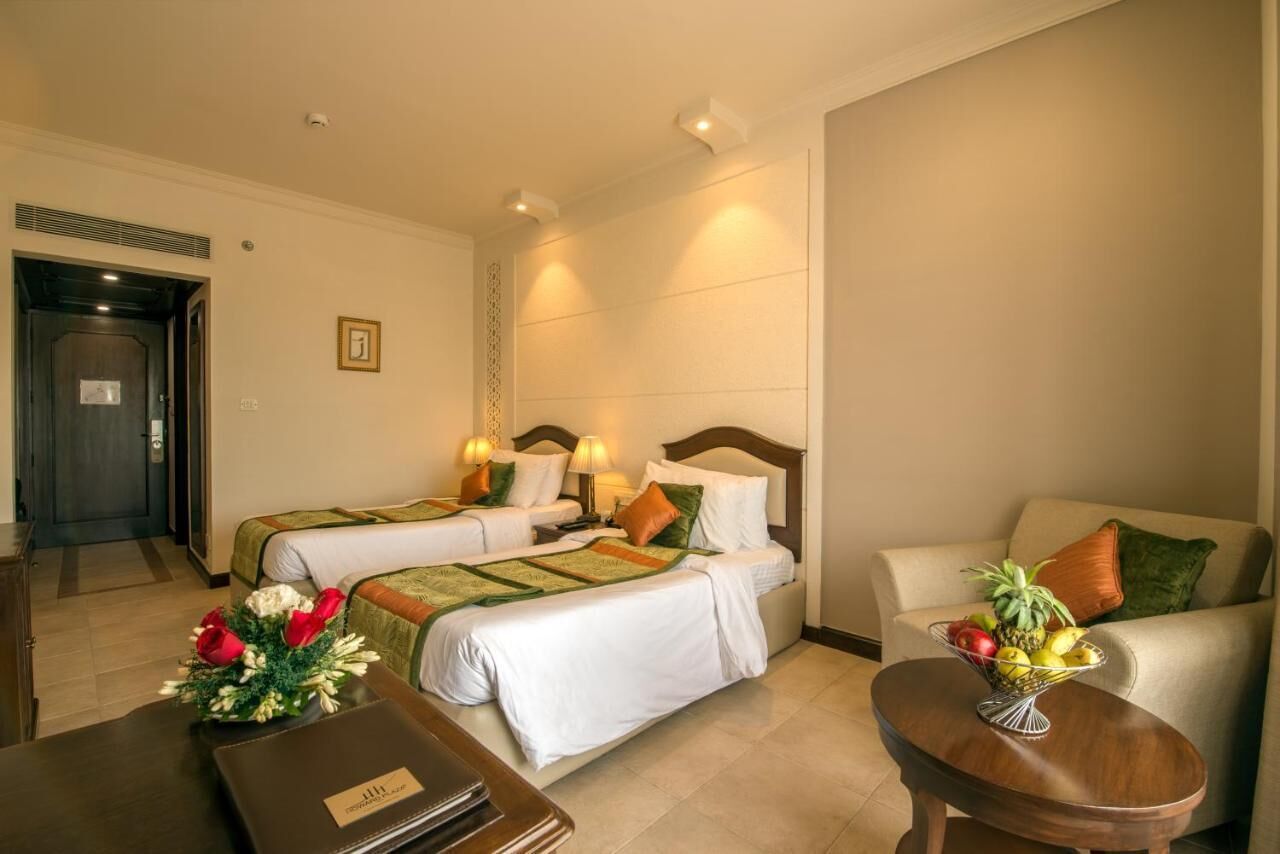 A hotel room with two beds, a sofa, a small table with a fruit bowl, and a door.