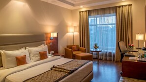 A hotel room with a large bed, two bedside tables, a desk, a chair, a small table with a lamp, and a window with curtains. - Howard Plaza The Fern, Agra (Agra)