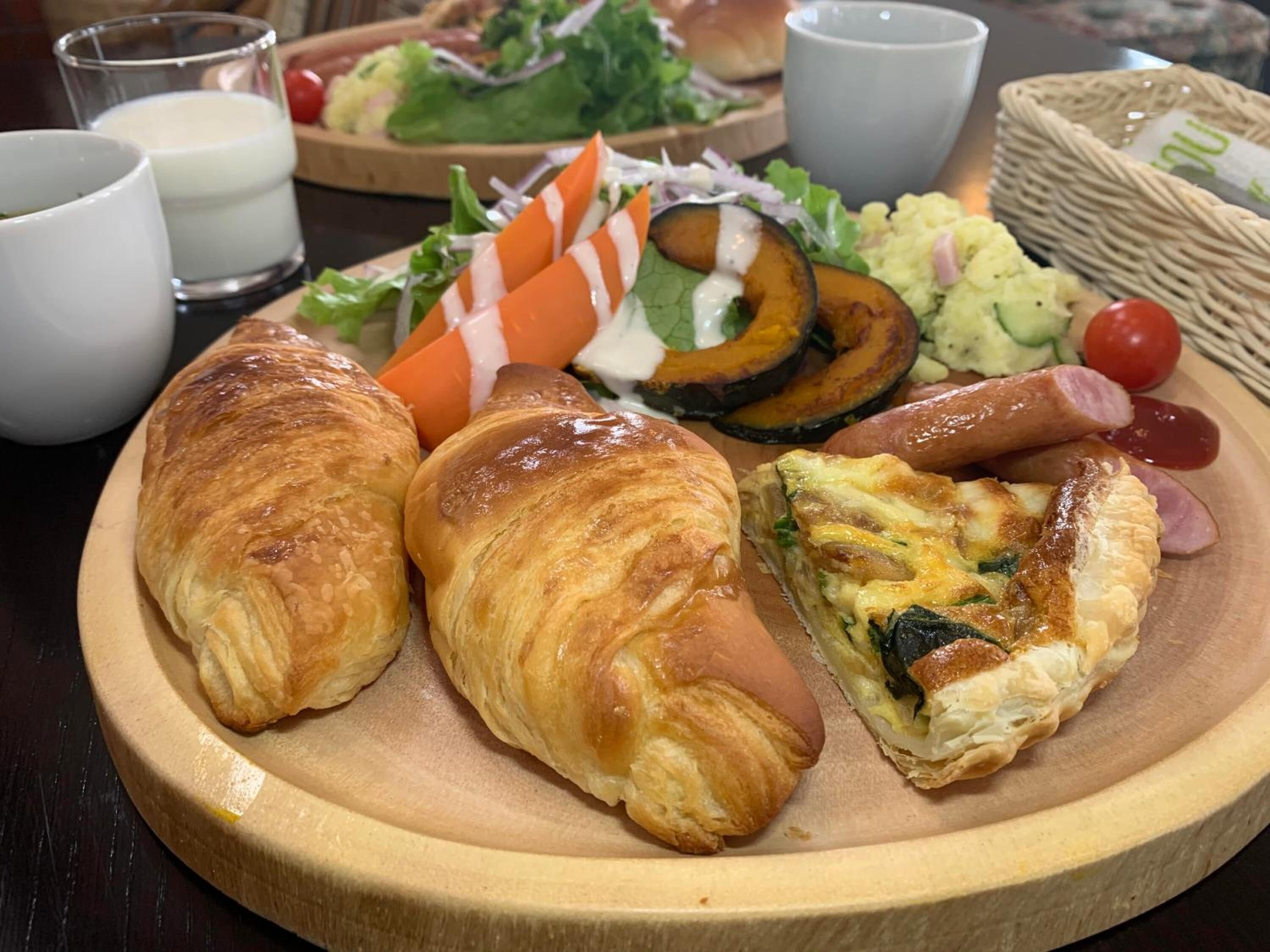 A wooden plate with croissants, a quiche, grilled vegetables, and a side salad.