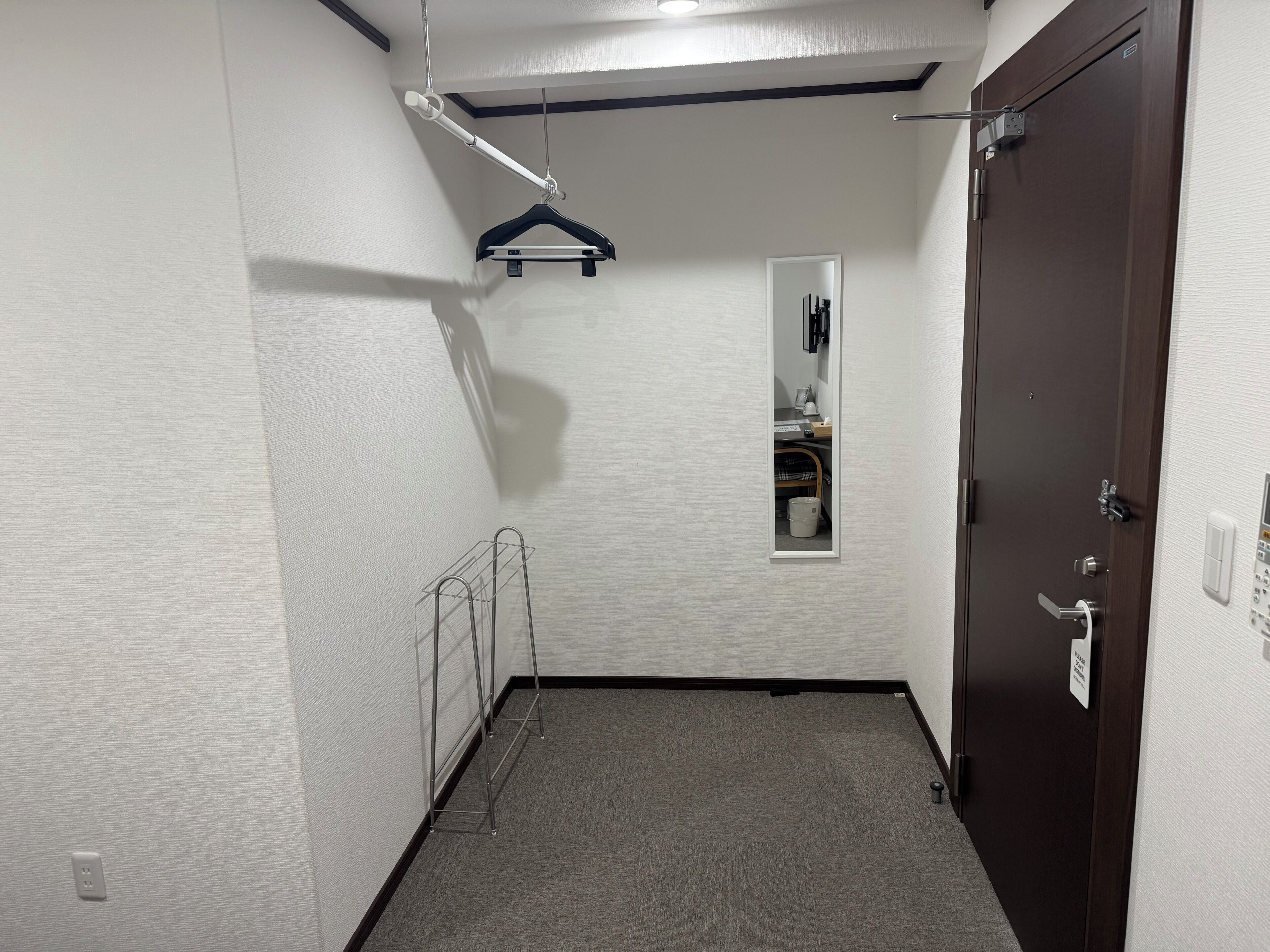 Quarto de hotel com porta de madeira escura, parede branca com espelho, cabideiro suspenso e arara de metal.