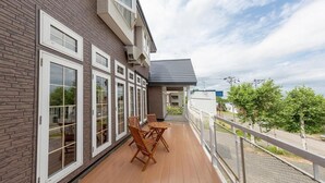 A balcony with a table and chairs, a view of the street, and a building with large windows. - Pension Lavender (Furano)