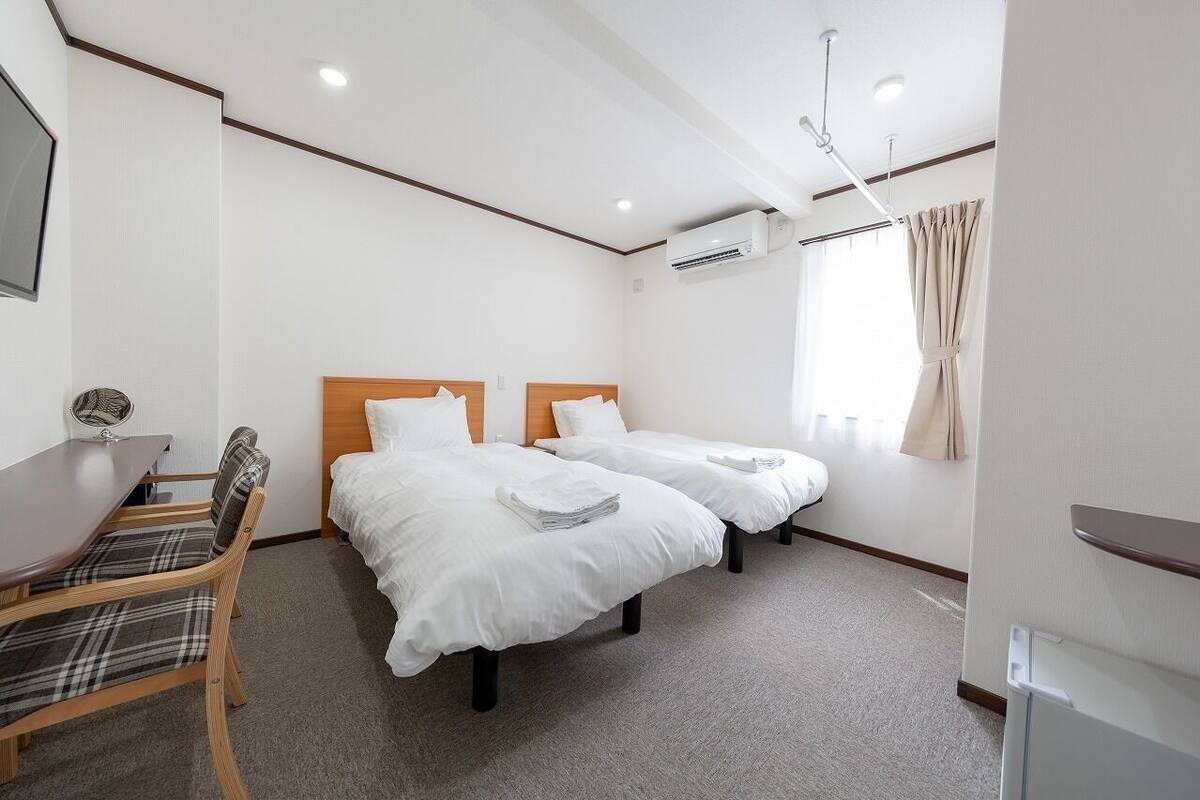 A hotel room with two beds, a desk, a chair, an air conditioning unit, and a window with curtains.