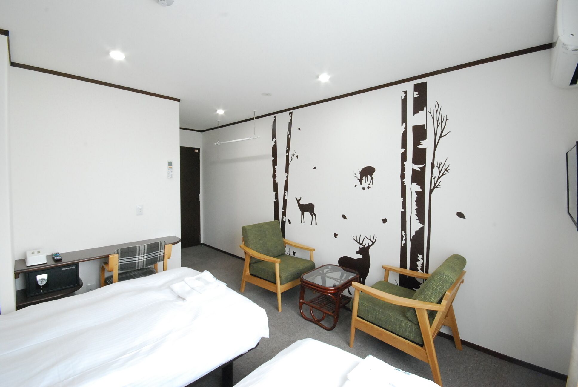 A hotel room with a bed, two green armchairs, a small table, and a wall with a forest decal.