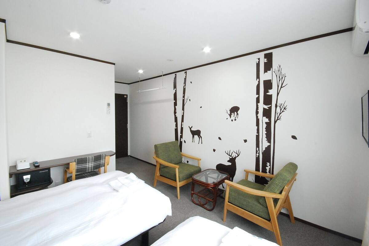 A hotel room with a bed, two green armchairs, a small table, and a wall with a forest decal.